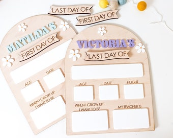 May include: Two wooden signs with arched tops, decorated with white flowers. The signs are for "Matilda's First Day of" and "Victoria's Last Day of". Each sign has spaces for recording age, date, height, and other information.