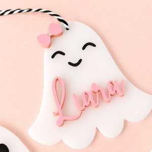 May include: White ghost-shaped ornament with a pink bow and the name "Liara" written in pink letters.