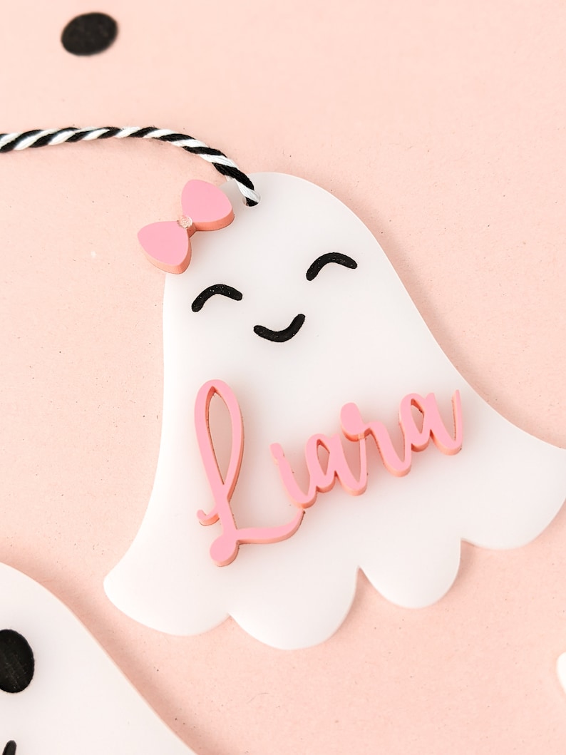May include: White ghost-shaped ornament with a pink bow and the name "Liara" written in pink letters.