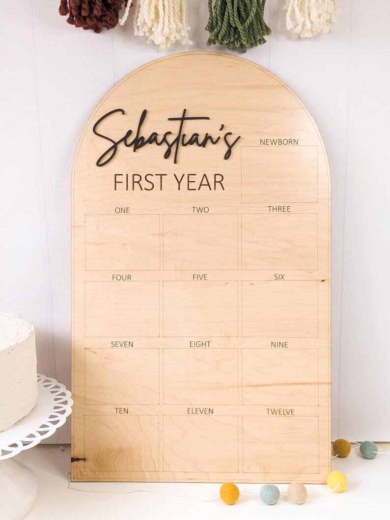 May include: A wooden milestone board with an arched top. The board is engraved with the text "Sebastian's FIRST YEAR" and numbered boxes for each month. The word "NEWBORN" is also included. Decorative elements include a cake and colorful felt balls.