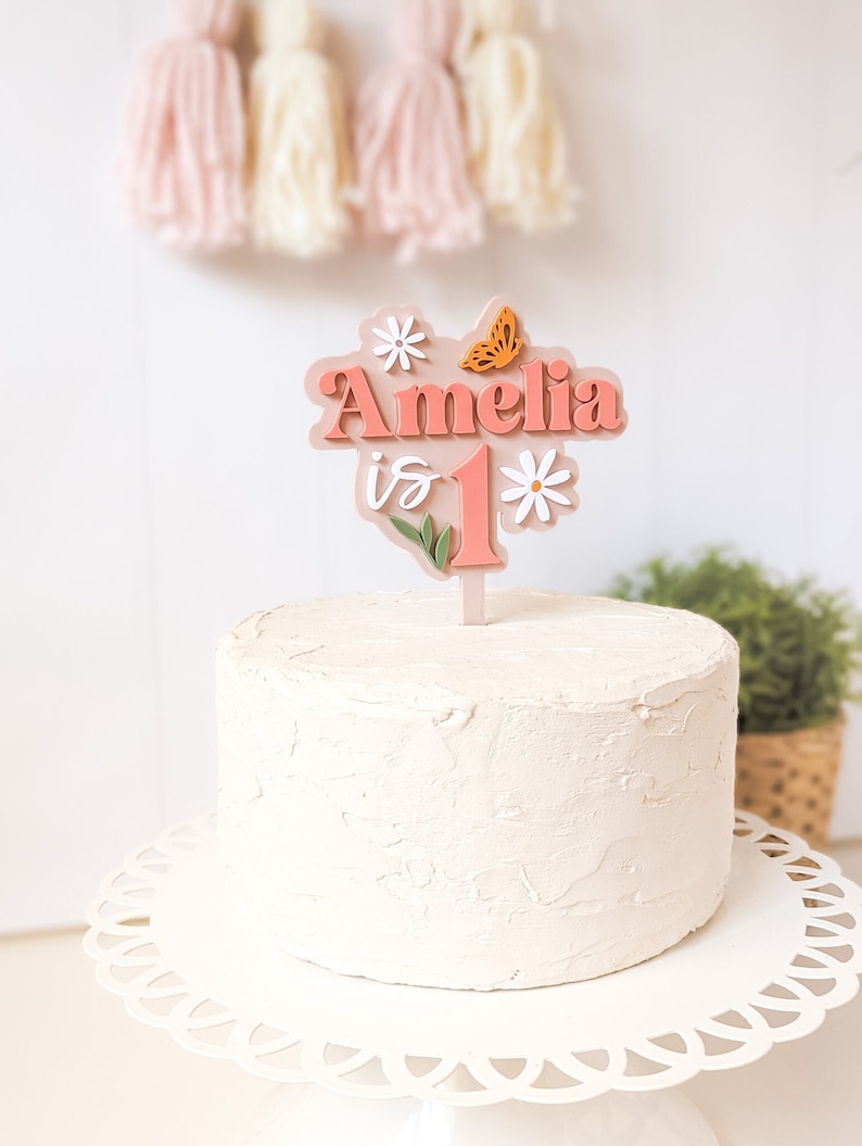 May include: A pink and white cake topper with the name Amelia and the number 1, decorated with flowers and a butterfly. The topper is on a white cake.
