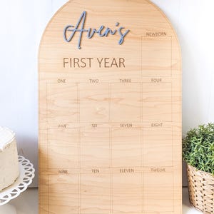 May include: Wooden arched milestone board with the name "Aven's" in blue script. The board is engraved with "FIRST YEAR" and boxes for each month, from newborn to twelve months. A decorative item for baby's first year.