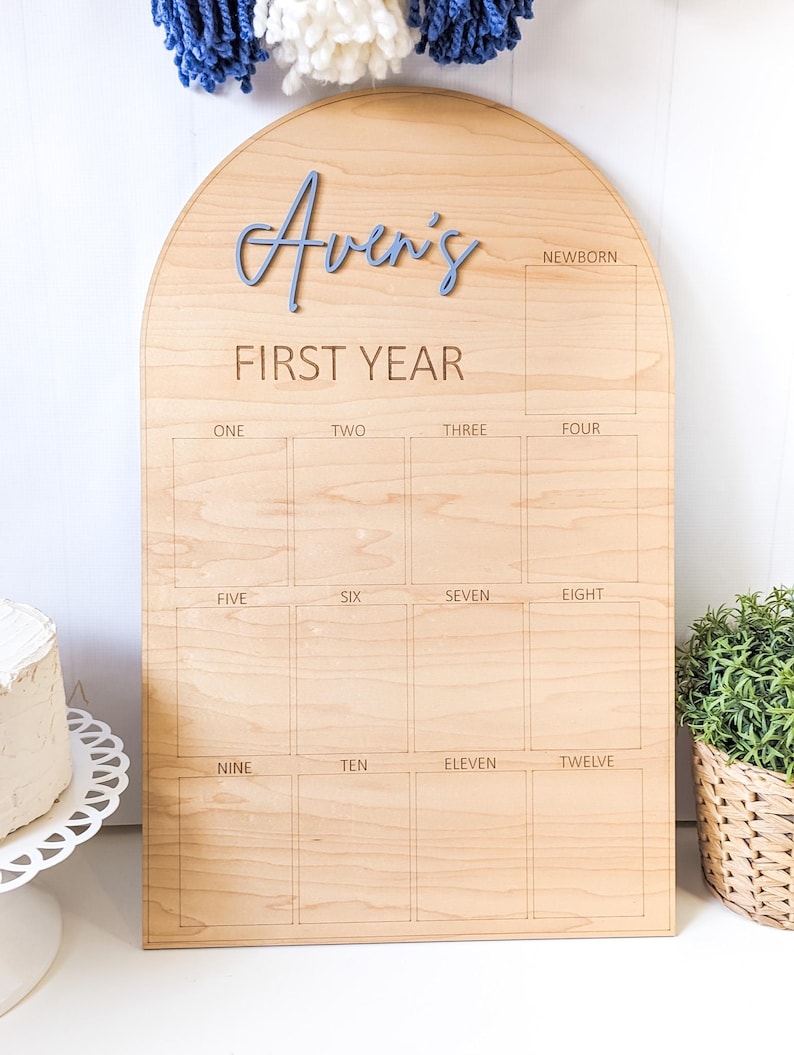 May include: Wooden arched milestone board with the name "Aven's" in blue script. The board is engraved with "FIRST YEAR" and boxes for each month, from newborn to twelve months. A decorative item for baby's first year.