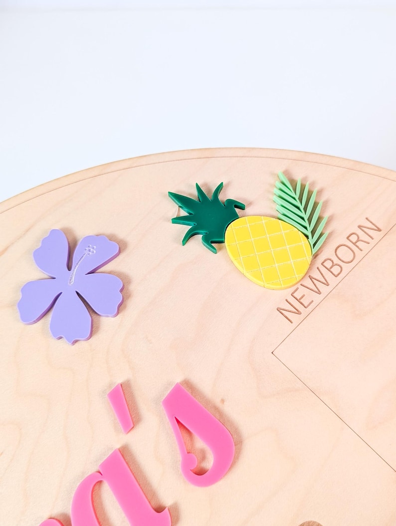 May include: A circular wooden plaque featuring a light wood grain finish. The plaque is decorated with a purple acrylic hibiscus flower, a yellow pineapple with green leaves, and the word NEWBORN engraved in capital letters. Pink acrylic letters are visible at the bottom.