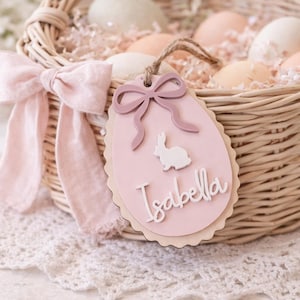 May include: A wicker Easter basket filled with eggs and decorative paper. A pink bow decorates the handle. A pink egg-shaped tag with a bunny and the name "Isabella" hangs from the basket.