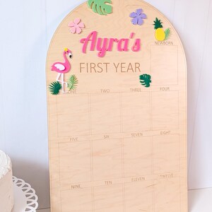 May include: An arched wooden milestone board featuring the text Ayra's FIRST YEAR. The board includes a grid for photos labeled from newborn to twelve months, decorated with colorful 3D acrylic accents including a pink flamingo, pineapple, and leaves.