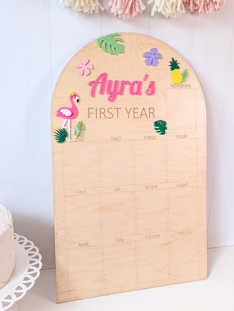 May include: An arched wooden milestone board featuring the text Ayra's FIRST YEAR. The board includes a grid for photos labeled from newborn to twelve months, decorated with colorful 3D acrylic accents including a pink flamingo, pineapple, and leaves.