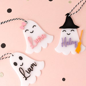 May include: Three white ghost-shaped ornaments with black eyes and mouths. The ghosts have strings attached to them. The ghosts are decorated with the names "Liam", "Laura", and "Haley".