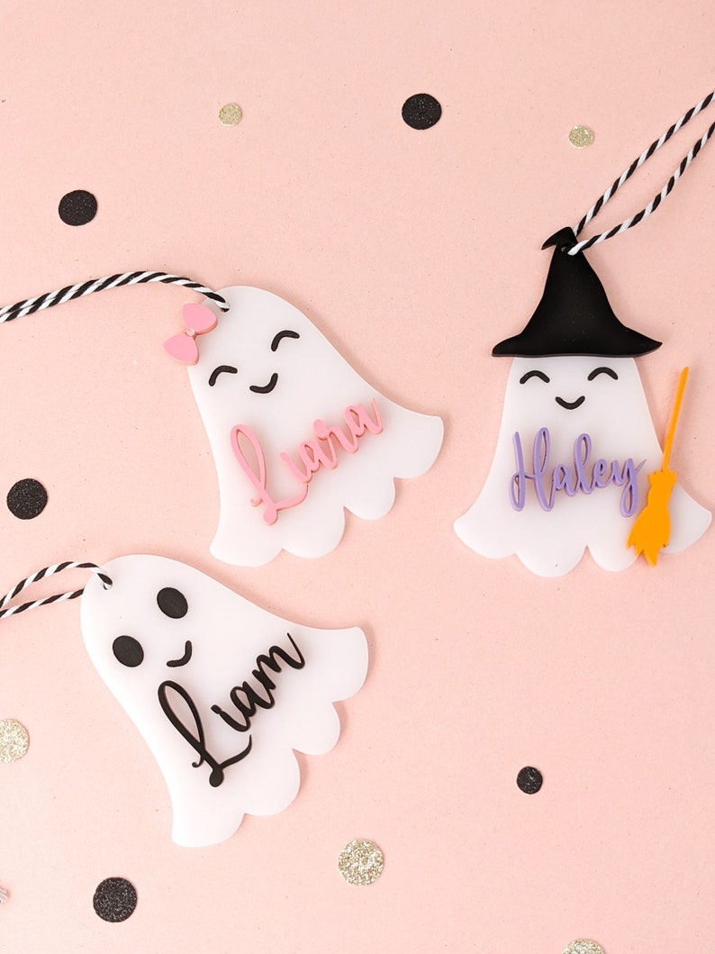 May include: Three white ghost-shaped ornaments with black eyes and mouths. The ghosts have strings attached to them. The ghosts are decorated with the names "Liam", "Laura", and "Haley".