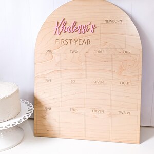 May include: A wooden milestone board with an arched top. The board is light brown with the name "Khalesst's" in pink, above "FIRST YEAR". It has boxes for each month, from newborn to twelve months. A cake sits on a white cake stand.