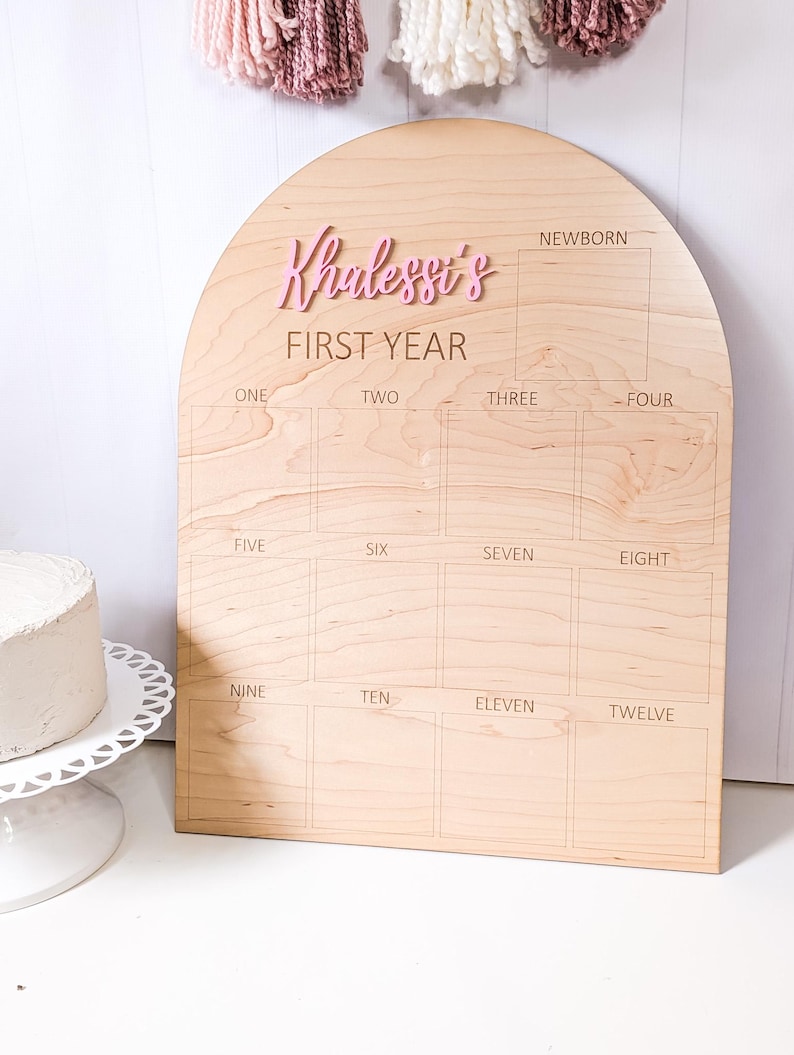 May include: A wooden milestone board with an arched top. The board is light brown with the name "Khalesst's" in pink, above "FIRST YEAR". It has boxes for each month, from newborn to twelve months. A cake sits on a white cake stand.