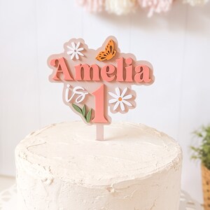 May include: A pink cake topper with the name Amelia and the number 1, decorated with flowers and a butterfly. The topper is on a white frosted cake.