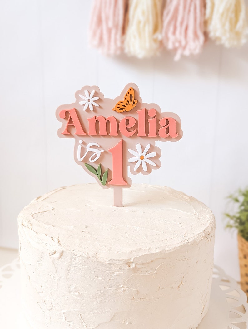 May include: A pink cake topper with the name Amelia and the number 1, decorated with flowers and a butterfly. The topper is on a white frosted cake.
