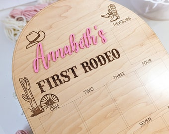 May include: A wooden milestone board with the text "Annabeth's FIRST RODEO" in pink and brown. The board features engraved cowboy-themed illustrations and boxes for tracking milestones. A cake and yarn are in the background.