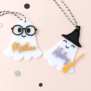 May include: Two white ghost-shaped Halloween gift tags with black and white twine. One ghost has black glasses and the name "Mattheo" in gold lettering. The other ghost has a black witch hat and the name "Haley" in purple lettering with a gold broom.