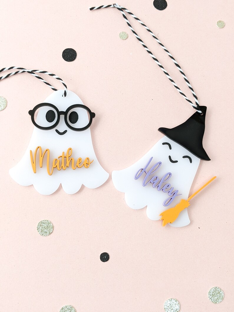 May include: Two white ghost-shaped Halloween gift tags with black and white twine. One ghost has black glasses and the name "Mattheo" in gold lettering. The other ghost has a black witch hat and the name "Haley" in purple lettering with a gold broom.