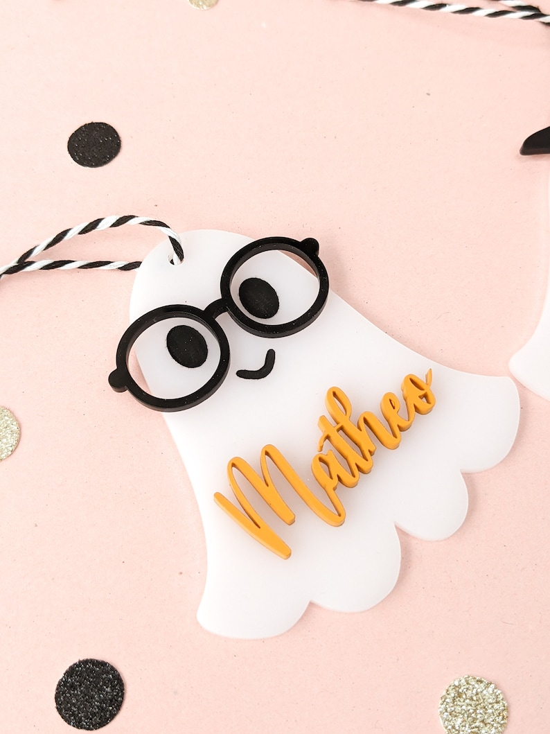 May include: White ghost-shaped gift tag with black glasses and a smiling face. The name "Mathew" is written in orange on the tag.