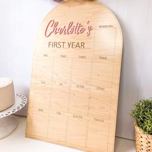May include: Wooden milestone board with an arched top. The board is light brown with the text "Charlotte's FIRST YEAR" in pink. It has spaces for each month, from newborn to twelve months. Decorative yarn tassels hang above.