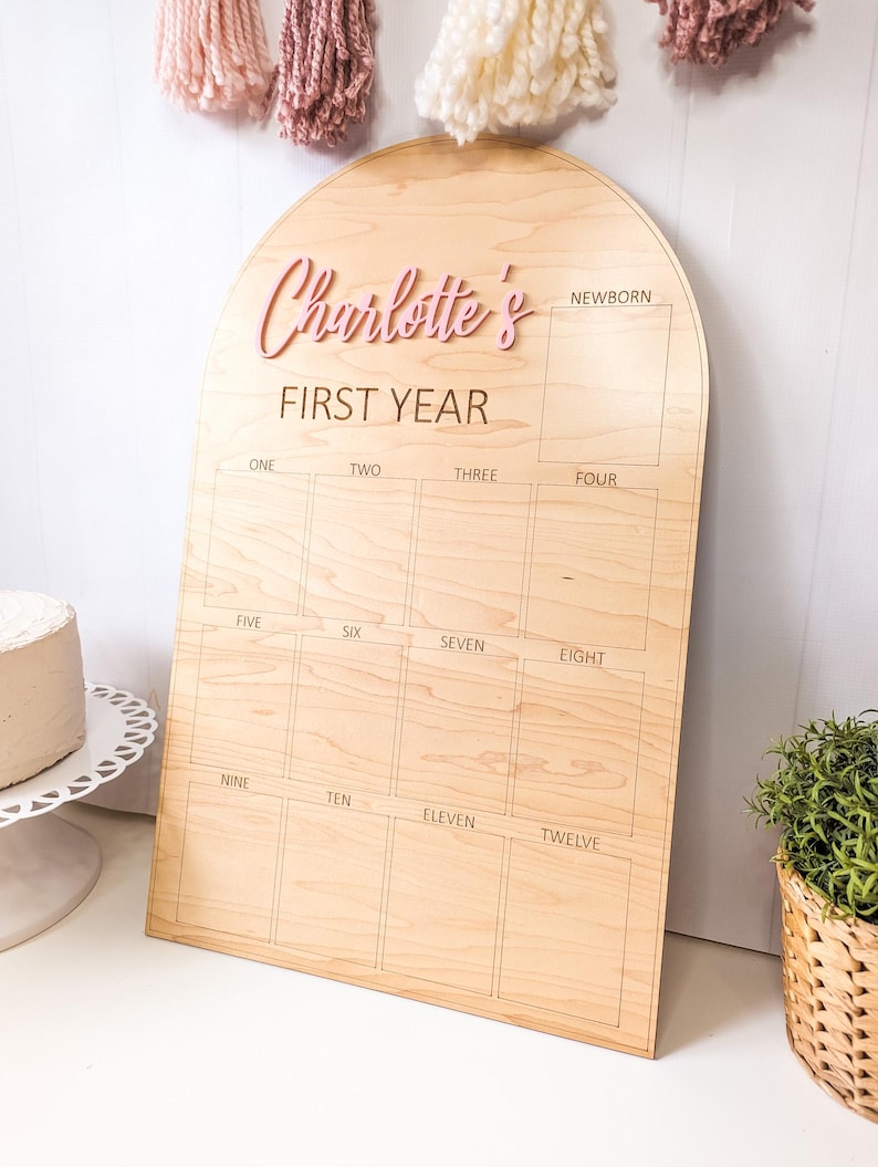 May include: Wooden milestone board with an arched top. The board is light brown with the text "Charlotte's FIRST YEAR" in pink. It has spaces for each month, from newborn to twelve months. Decorative yarn tassels hang above.