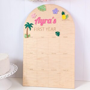 May include: An arched wooden milestone board featuring the text Ayra's FIRST YEAR. The board includes a grid for photos labeled from NEWBORN to TWELVE, decorated with colorful wooden cutouts of a palm tree, flowers, a pineapple, and leaves.