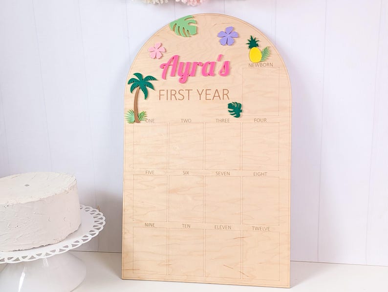 May include: An arched wooden milestone board featuring the text Ayra's FIRST YEAR. The board includes a grid for photos labeled from NEWBORN to TWELVE, decorated with colorful wooden cutouts of a palm tree, flowers, a pineapple, and leaves.