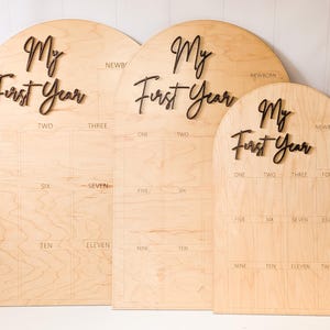 May include: Three wooden "My First Year" milestone boards. Each board has a light wood finish with dark brown lettering. The boards are arched at the top and have spaces for monthly photos.