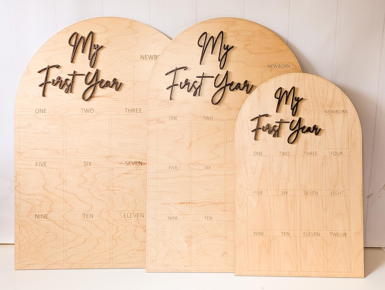 May include: Three wooden "My First Year" milestone boards. Each board has a light wood finish with dark brown lettering. The boards are arched at the top and have spaces for monthly photos.