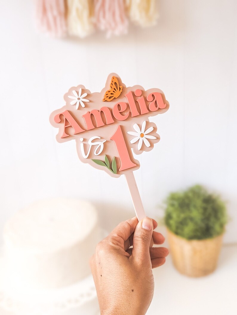 May include: Pink and white cake topper with the name Amelia and the number 1, decorated with flowers and a butterfly. The topper says "Amelia is 1".