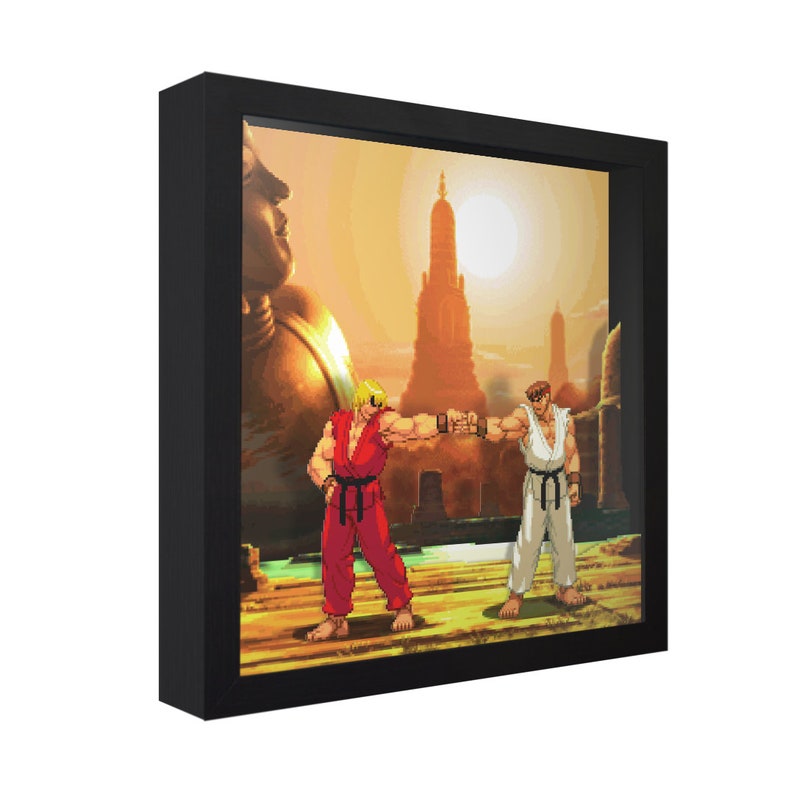 May include: A framed pixel art print of two characters from a video game, one in red and one in white, facing each other with fists raised in a fighting pose. The background is a sunset scene with a large golden statue and a temple in the distance.