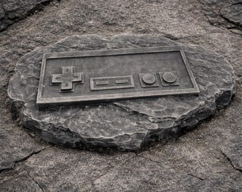 May include: A gray stone carving of a classic video game controller, featuring a directional pad, two rectangular buttons, and two circular buttons. The controller is set into a rough-hewn stone, creating a retro gaming aesthetic.