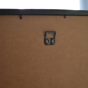 May include: The back of a black picture frame with a brown cardboard backing. A black plastic hanging hook is attached to the back of the frame.