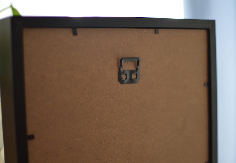 May include: The back of a black picture frame with a brown cardboard backing. A black plastic hanging hook is attached to the back of the frame.