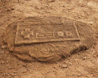 May include: A brown, textured stone carving of a classic video game controller. The controller is rectangular with a directional pad, buttons, and a textured surface. The carving is set in a sandy environment.