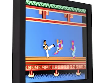 May include: A framed pixel art print of the classic video game 'Karateka'. The print features a scene from the game, with the main character in a fighting stance, facing off against two opponents. The print is set against a blue background with a red and white platform in the foreground.