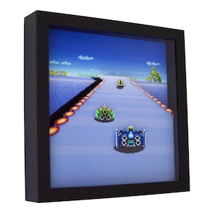 May include: A framed pixel art print of a retro video game scene. The image shows three spaceships racing on a blue and white track. The spaceships are green, blue, and red. The print is in a black frame.
