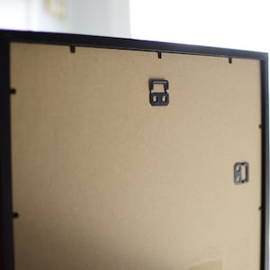 May include: Black picture frame with a brown cardboard backing. The frame has two metal hanging hooks on the back.