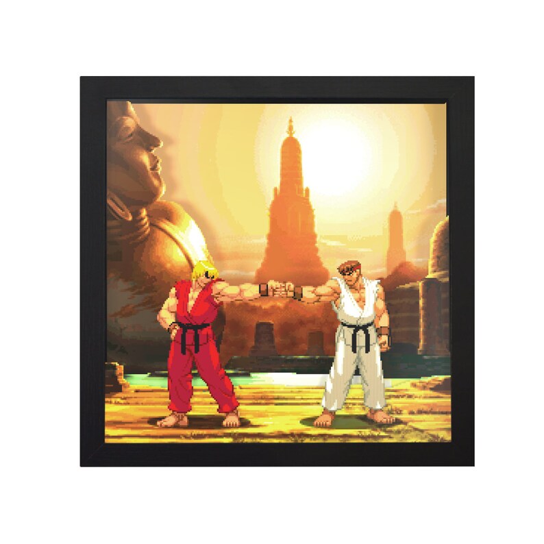 May include: A framed pixel art print of two video game characters, Ryu and Ken, facing each other with fists raised in a fighting pose. The background is a sunset scene with a large golden Buddha statue and a temple in the distance.