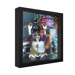 May include: A 3D lenticular art print in a black frame featuring a white, winged, cartoon character with a zipper on its belly. The character is wearing a crown and has red wings. The background is a colorful cityscape with neon signs.