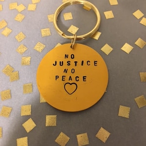 Puede incluir: Un llavero de oro con un dije redondo amarillo que dice "NO JUSTICE NO PEACE" con un símbolo de corazón.