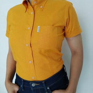 Peut inclure: Une chemise boutonn&eacute;e &agrave; manches courtes de couleur jaune avec une petite poche sur la poitrine gauche. La poche a une &eacute;tiquette blanche avec le texte "Japan Gear" dessus.