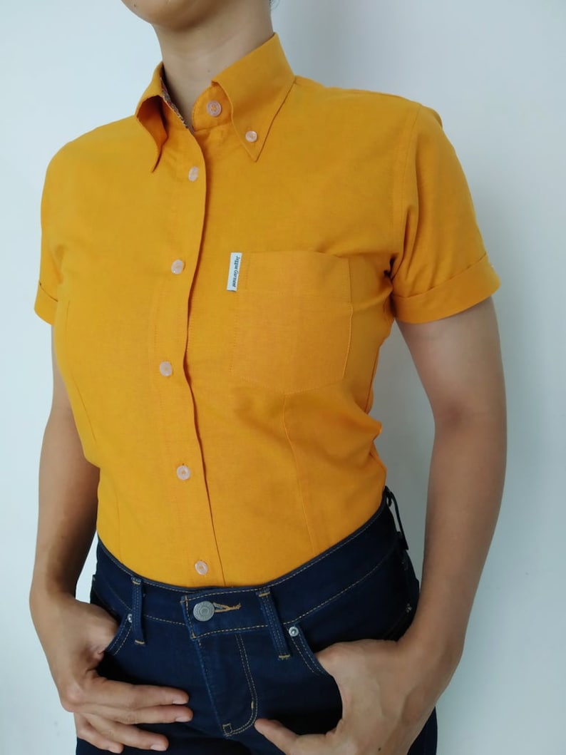 Peut inclure: Une chemise boutonn&eacute;e &agrave; manches courtes de couleur jaune avec une petite poche sur la poitrine gauche. La poche a une &eacute;tiquette blanche avec le texte "Japan Gear" dessus.
