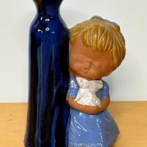 May include: A ceramic vase and figurine set. The vase is a tall, dark blue with a flared top. A figurine of a girl in a blue dress is leaning against the vase, holding a white object. The figurine has closed eyes and a serene expression.