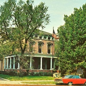 May include: A vintage postcard image of a large brick house with white columns and an American flag. A red car is parked in front of the house, and the scene is framed by lush green trees and a blue sky.
