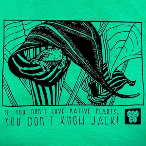 May include: A bright green t-shirt with a black graphic of a frog in a plant. The text "IF YOU DON'T LOVE NATIVE PLANTS, YOU DON'T KNOW JACK!" is printed below the graphic. The shirt also has a small tag with the brand name.