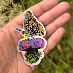 May include: A holographic sticker featuring a detailed butterfly with orange, black, and white patterns. The butterfly rests on a purple and green flower with the text "NATIVE PLANTS RULE!"
