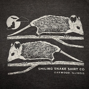 May include: Dark grey t-shirt with a white illustration of two opossums on branches. A full moon and cloud are in the upper left corner. The text "Smiling Snake Shirt Co Oakwood, Illinois" is printed below the opossums.