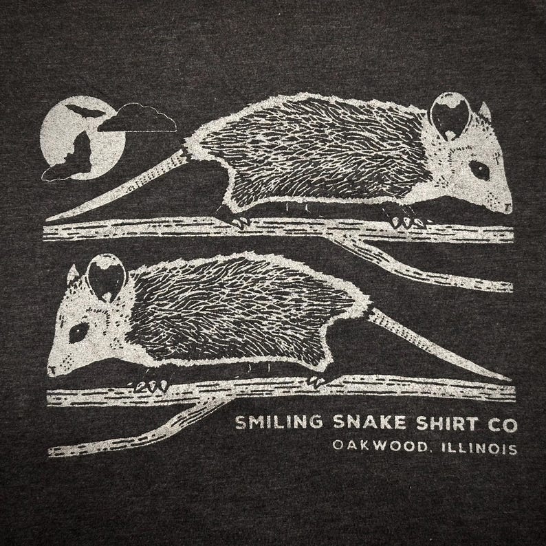 May include: Dark grey t-shirt with a white illustration of two opossums on branches. A full moon and cloud are in the upper left corner. The text "Smiling Snake Shirt Co Oakwood, Illinois" is printed below the opossums.