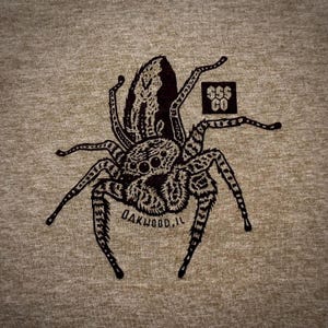 May include: A tan t-shirt featuring a detailed black illustration of a jumping spider. The spider design includes the text "OAKWOOD, IL" and a small square logo with the text "$$$ CO".