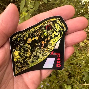 May include: A sticker featuring a detailed illustration of a turtle's head in yellow and black. The sticker has the text "XING GUARD" in red and white. The sticker is held in a hand.