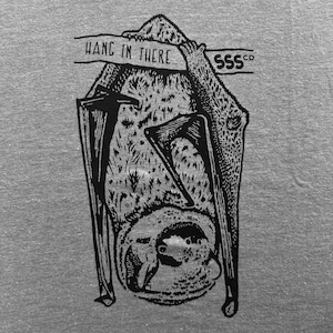 May include: Gray t-shirt featuring a black illustration of a bat hanging upside down. The bat is holding onto a branch with the words "HANG IN THERE" printed above it. The brand name "SSS co" is also printed on the branch.